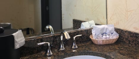 A bathroom with a granite countertop, a sink, a mirror, a towel dispenser, and a basket with toiletries.