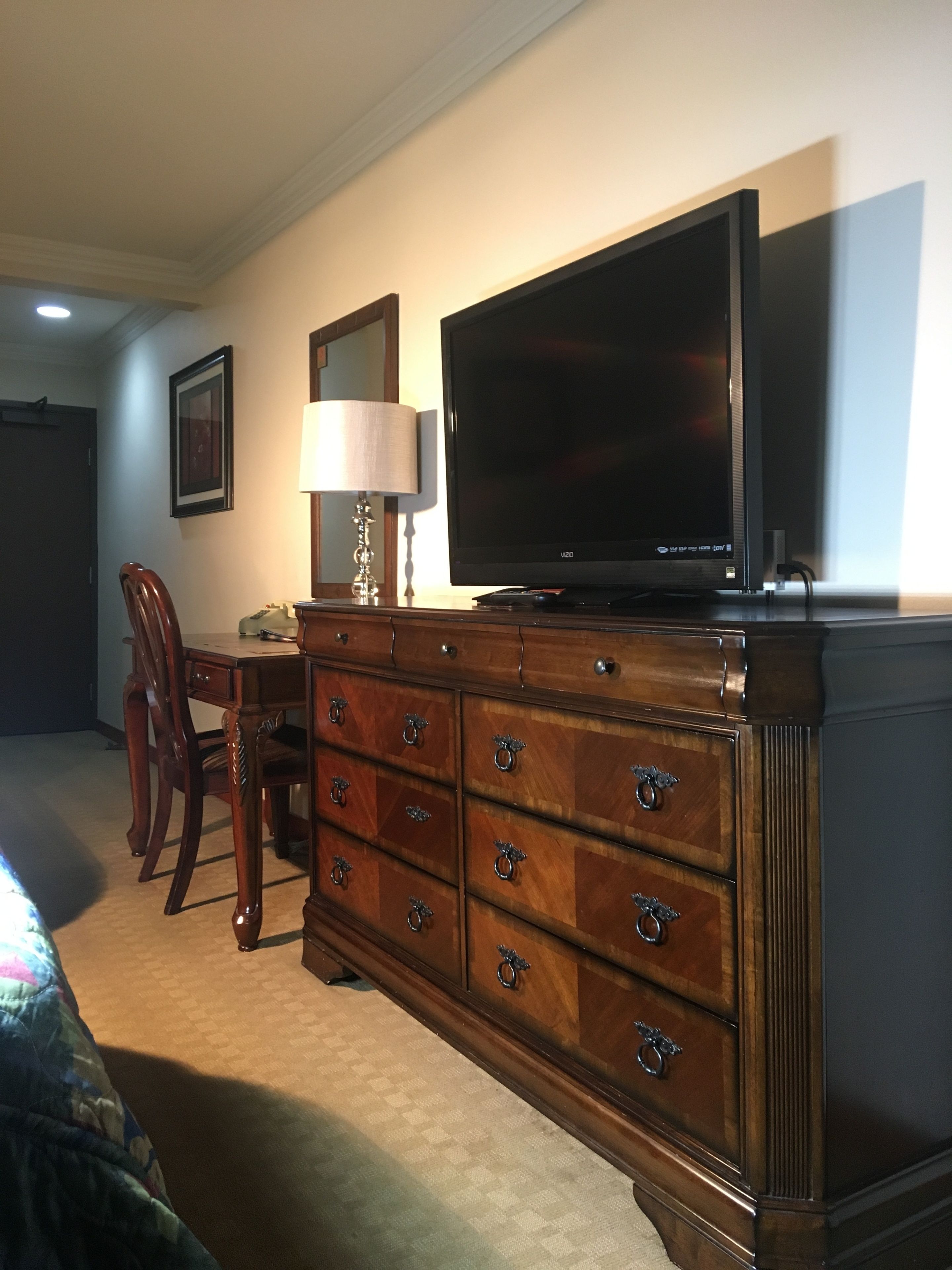 Une chambre d’hôtel avec une commode en bois, un téléviseur à écran plat, une lampe, un tableau encadré au mur et un coin repas comprenant une chaise et une table.