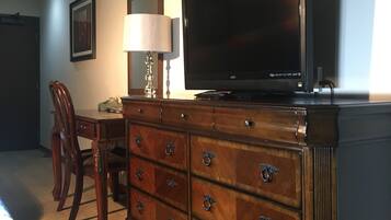 Une chambre d’hôtel avec une commode en bois, un téléviseur à écran plat, une lampe, un tableau encadré au mur et un coin repas comprenant une chaise et une table.