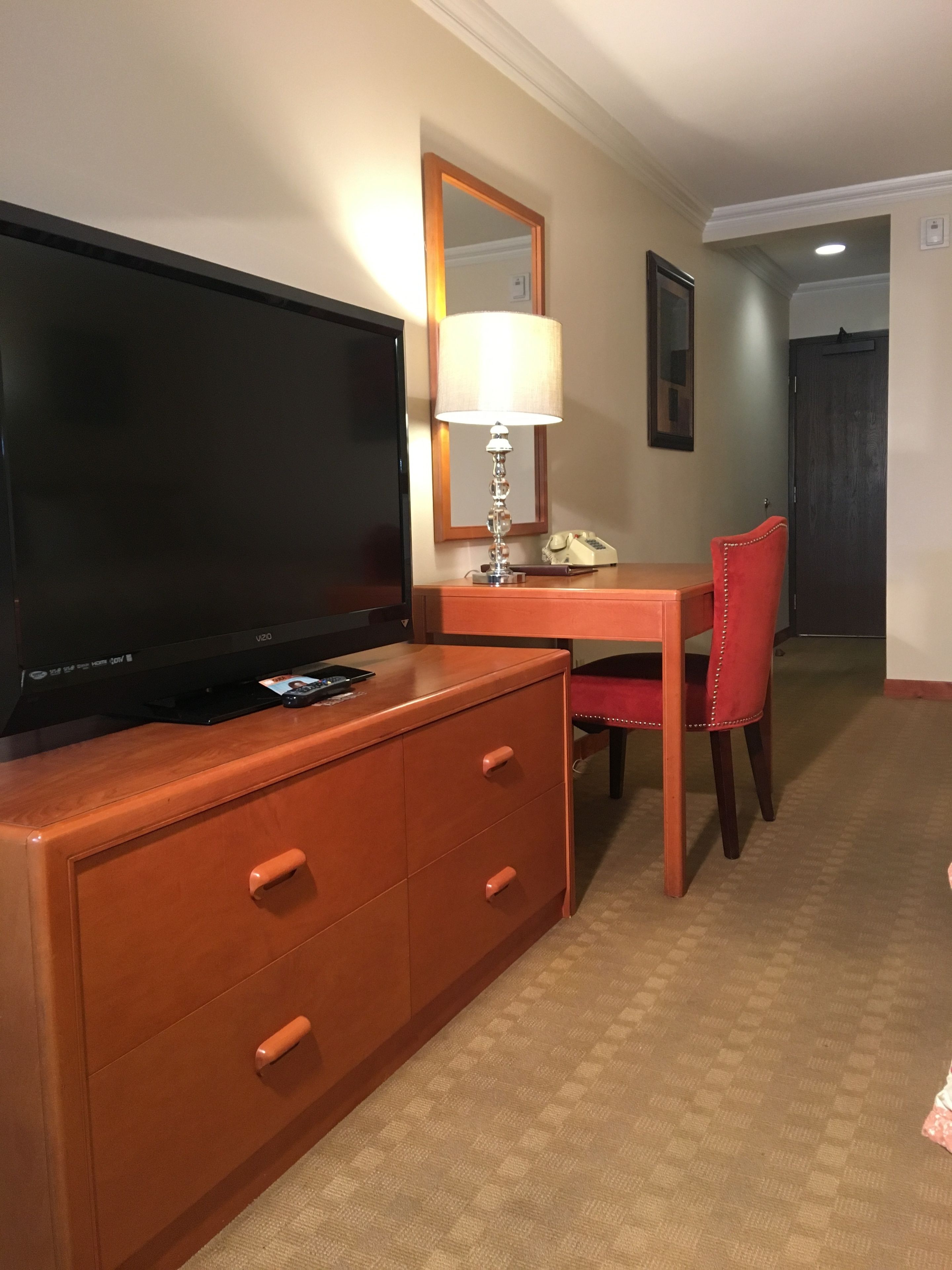 A hotel room with a TV on a wooden dresser, a desk with a lamp, a chair, and a mirror on the wall.