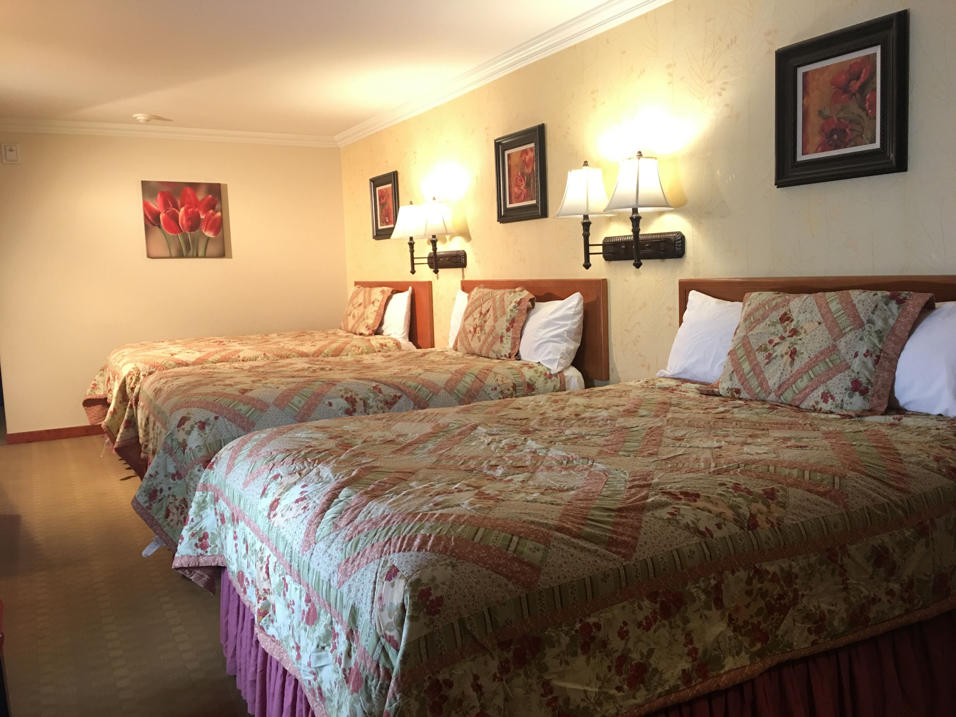 A hotel room with two beds, each with floral-patterned bedding and headboards. There are wall-mounted lamps and framed floral artwork above the beds.