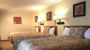A hotel room with two beds, each with floral-patterned bedding and headboards. There are wall-mounted lamps and framed floral artwork above the beds.