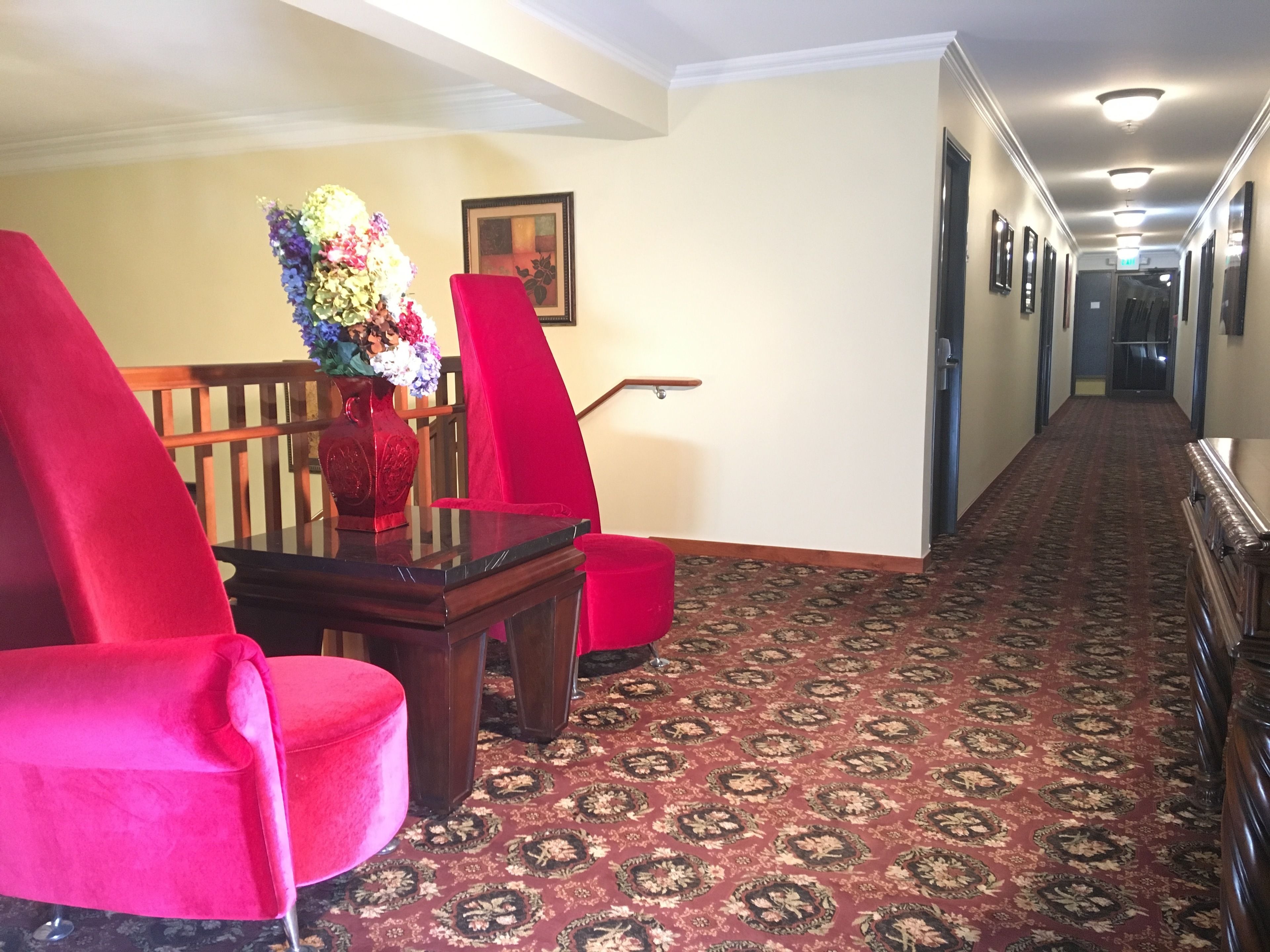A hallway with a carpeted floor, red chairs, a wooden table, and a vase of flowers.