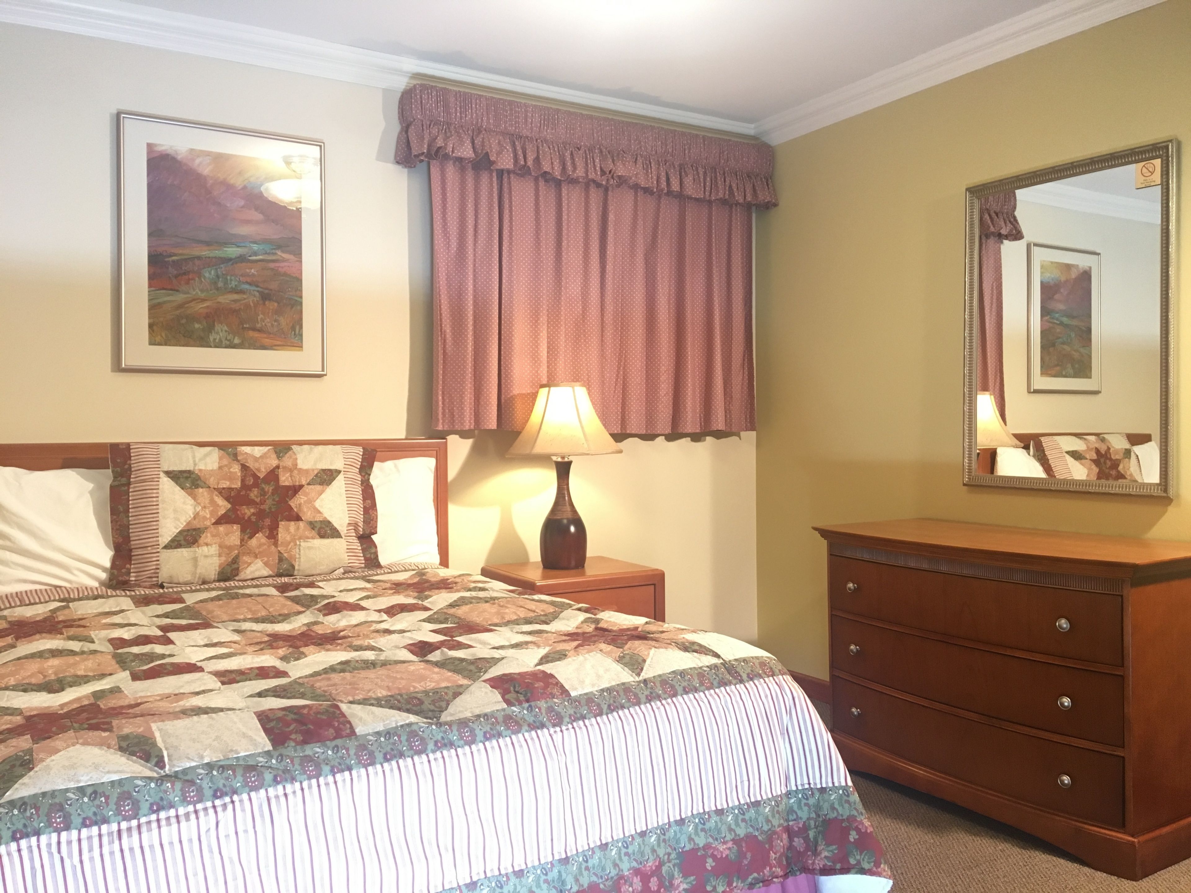 Une chambre d’hôtel avec un grand lit, une table de chevet avec une lampe, une commode, un tableau au mur et une fenêtre avec des rideaux.