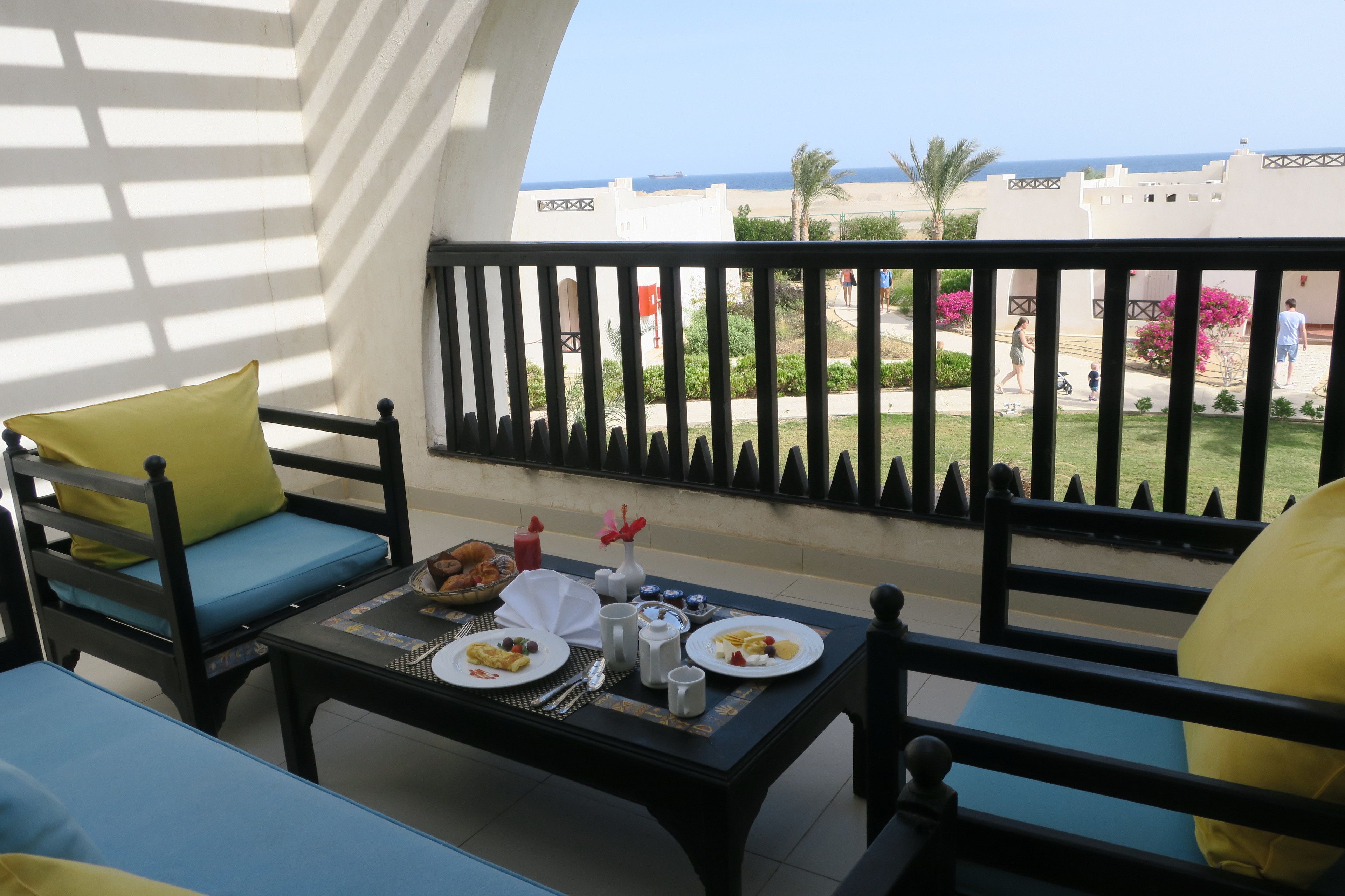 Varanda com mesa posta para café da manhã, vista para uma praia e palmeiras.