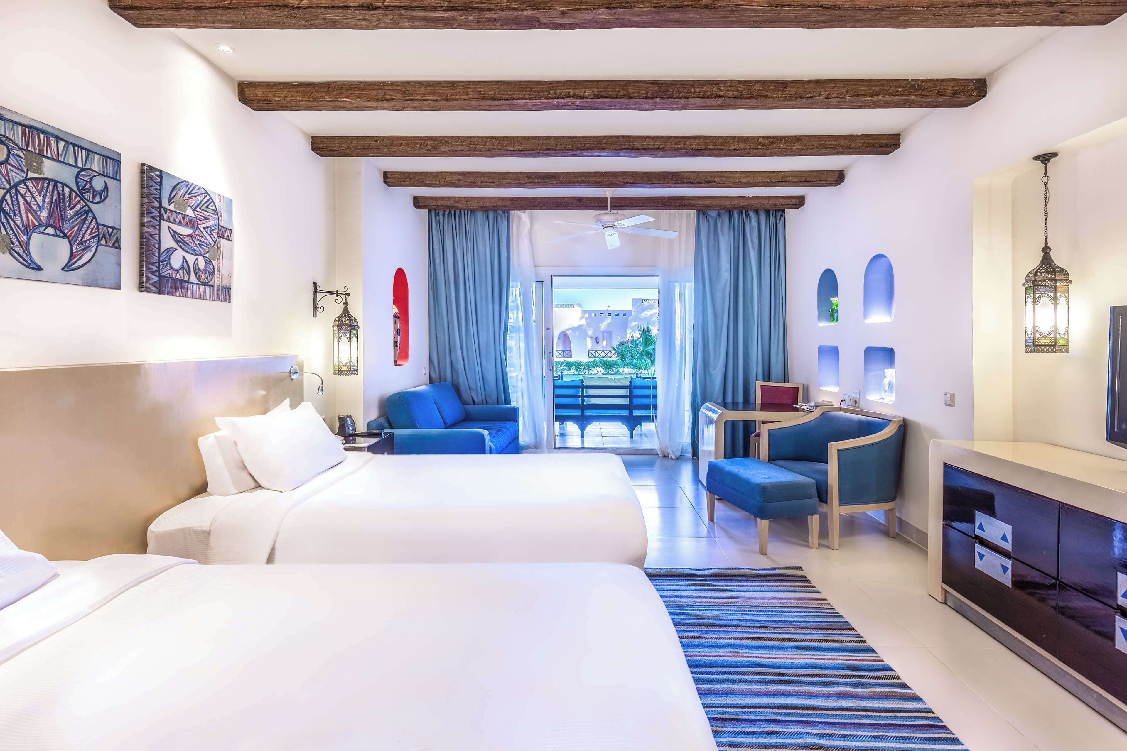 A hotel room with two beds, a desk, a chair, and a balcony with a view.