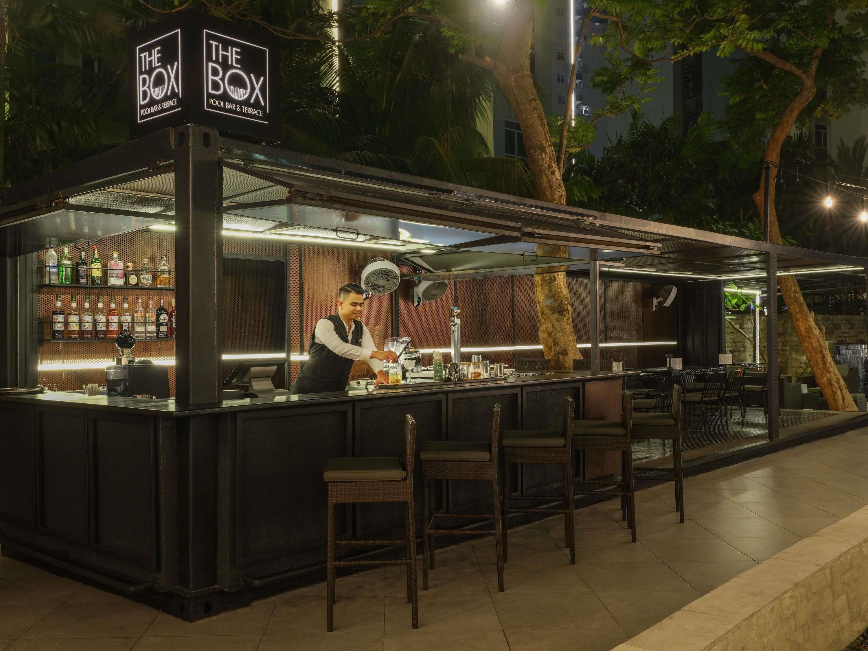 Zona de bar al aire libre con un bartender, una barra repleta de botellas y sillas.