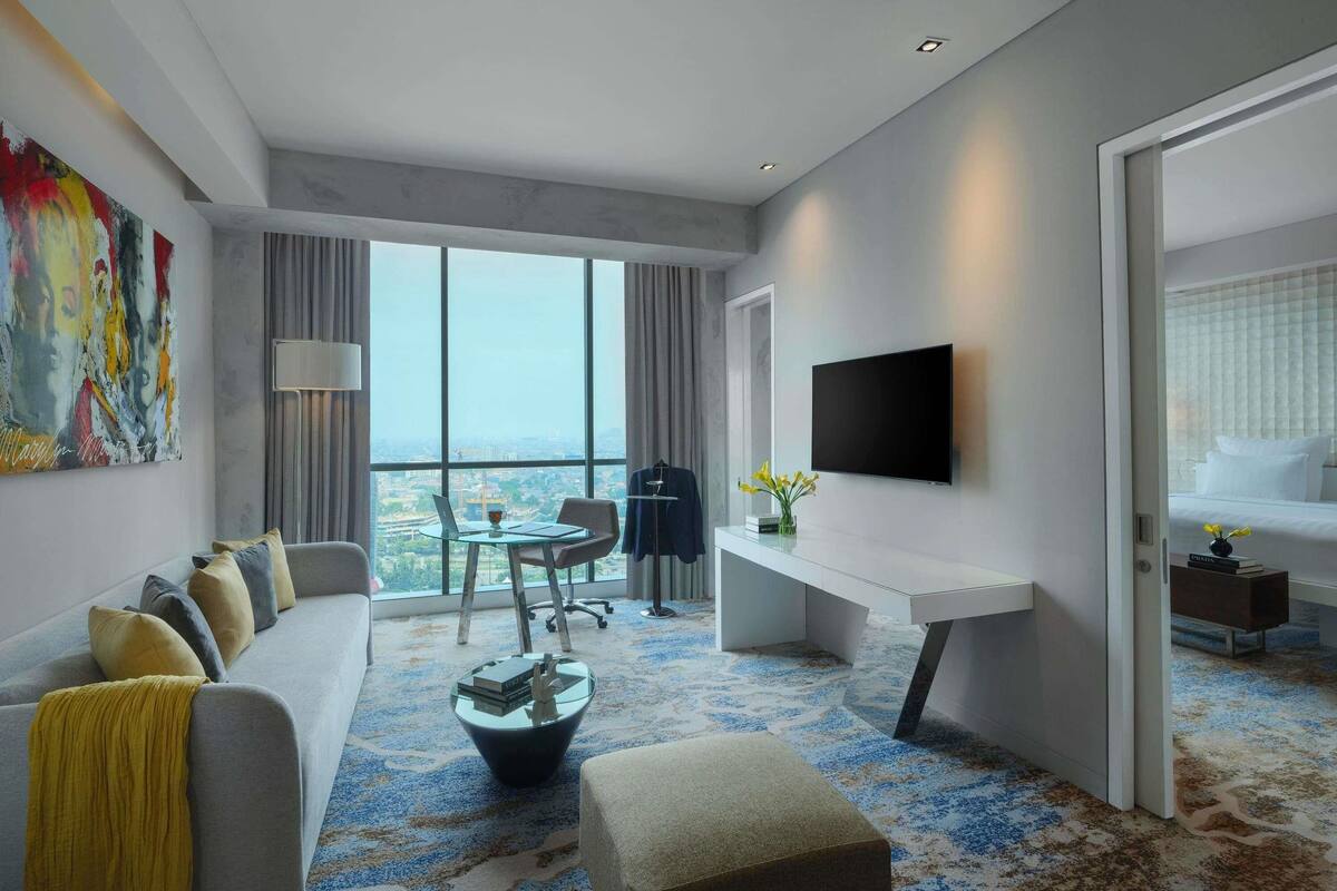 A modern hotel room with a sofa, a desk, a TV, and a large window offering a city view.