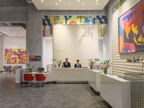 Un área de recepción moderna con mostrador, dos empleados, un cuadro abstracto grande y una zona de estar con sillas rojas.