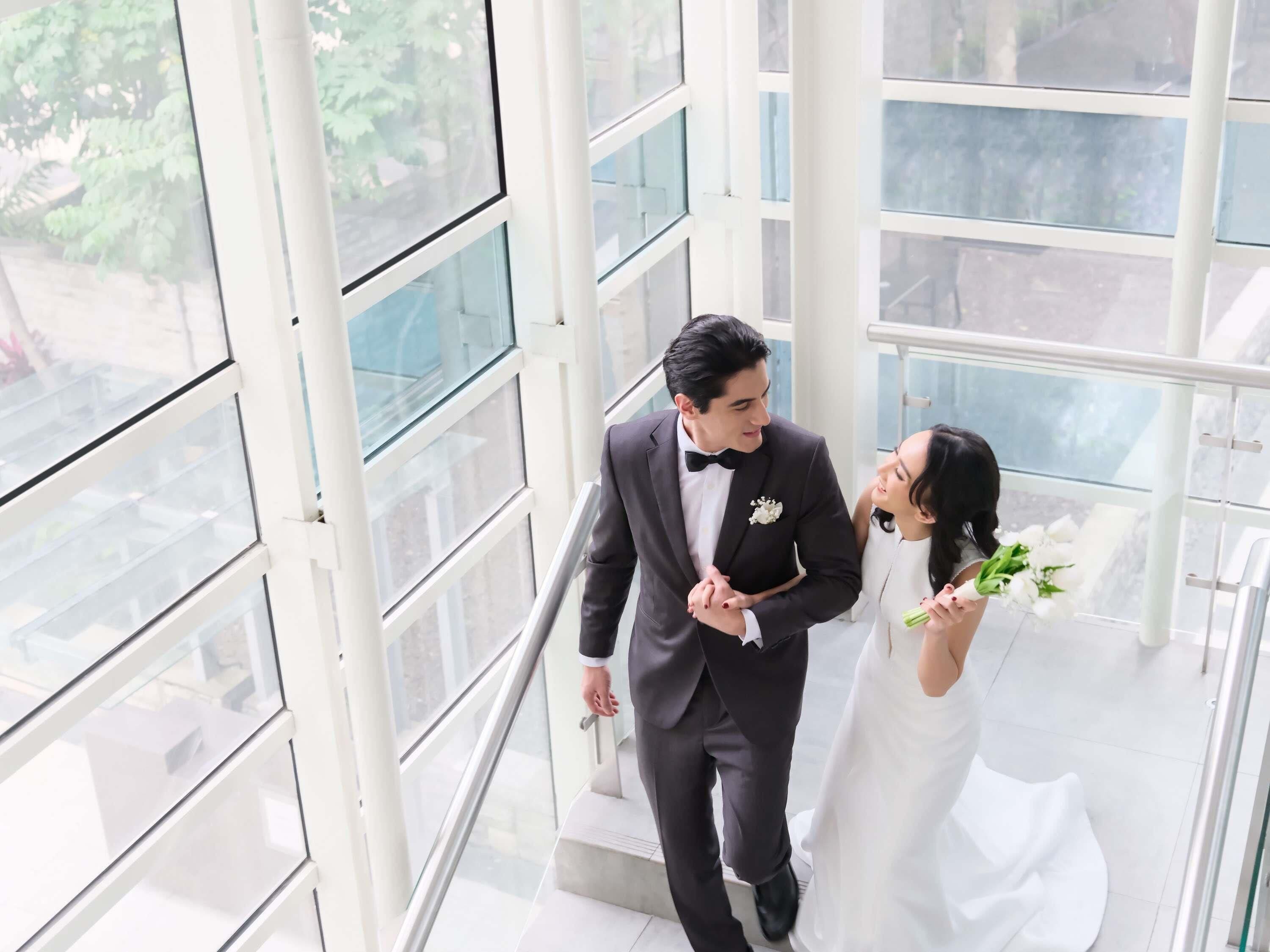 A couple in wedding attire on a staircase with large windows and a view of greenery outside.