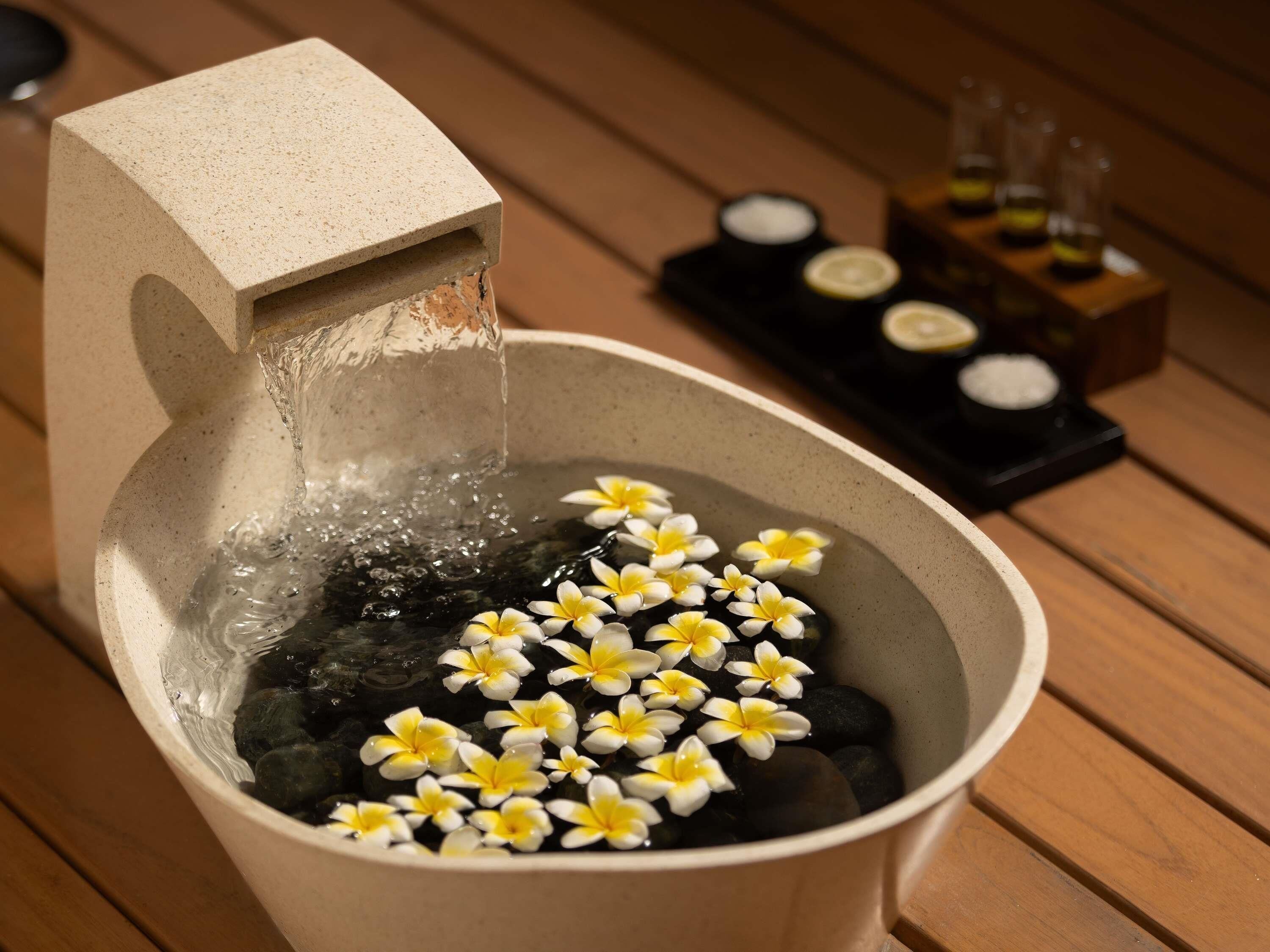 Un cuenco de piedra con agua que fluye desde un caño, rodeado de flores y piedras, con una bandeja de velas al fondo.