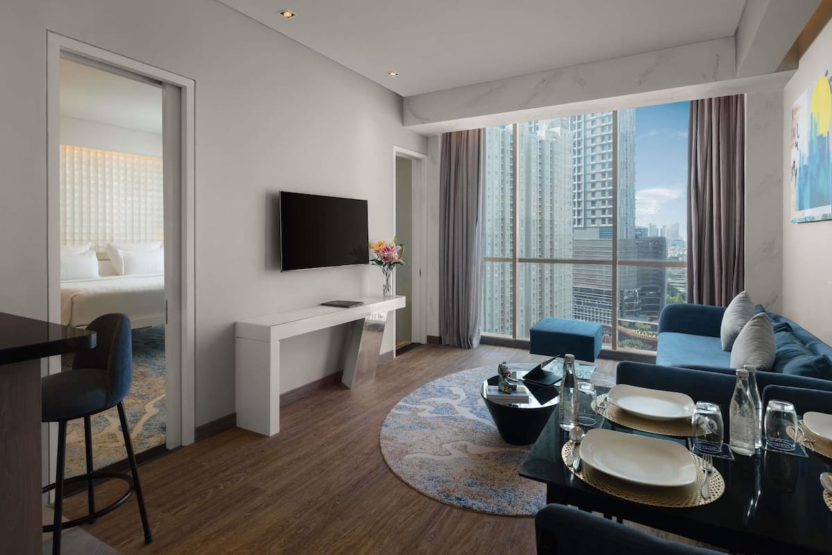 A modern hotel room with a flat-screen TV, a dining area with a round table, a sofa, and a large window offering a city view.