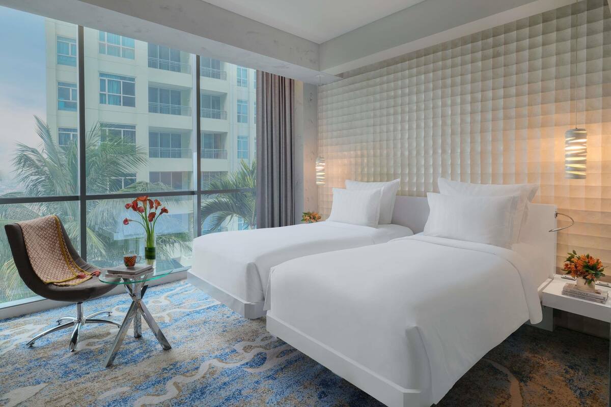 A hotel room with two beds, a chair, a small table, and a large window with a view of a building.