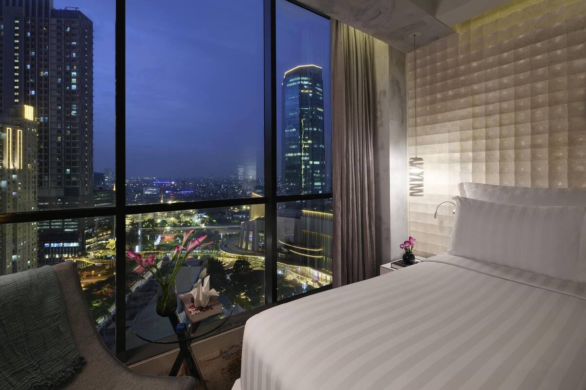 A hotel room with a large window offering a city view, a bed, a bedside table with a vase of flowers, and a modern light fixture.