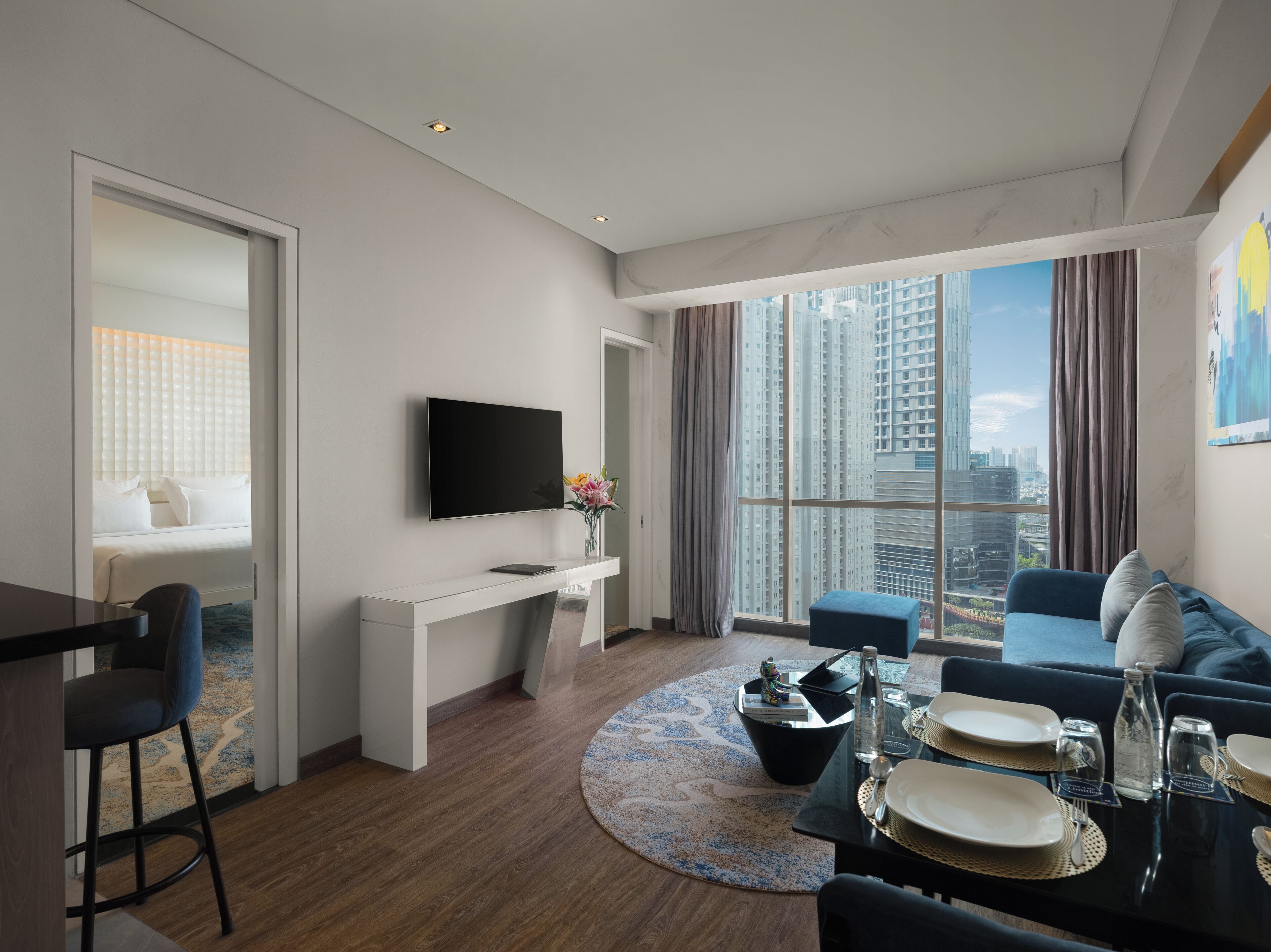 A modern hotel room with a flat-screen TV, a dining area with table set for two, a sofa, and a large window with a city view.
