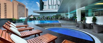 A rooftop pool area with wooden loungers, a hot tub, and a glass-enclosed bar area.