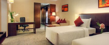 A hotel room with two beds, a desk with a chair, a lamp, and a painting on the wall.