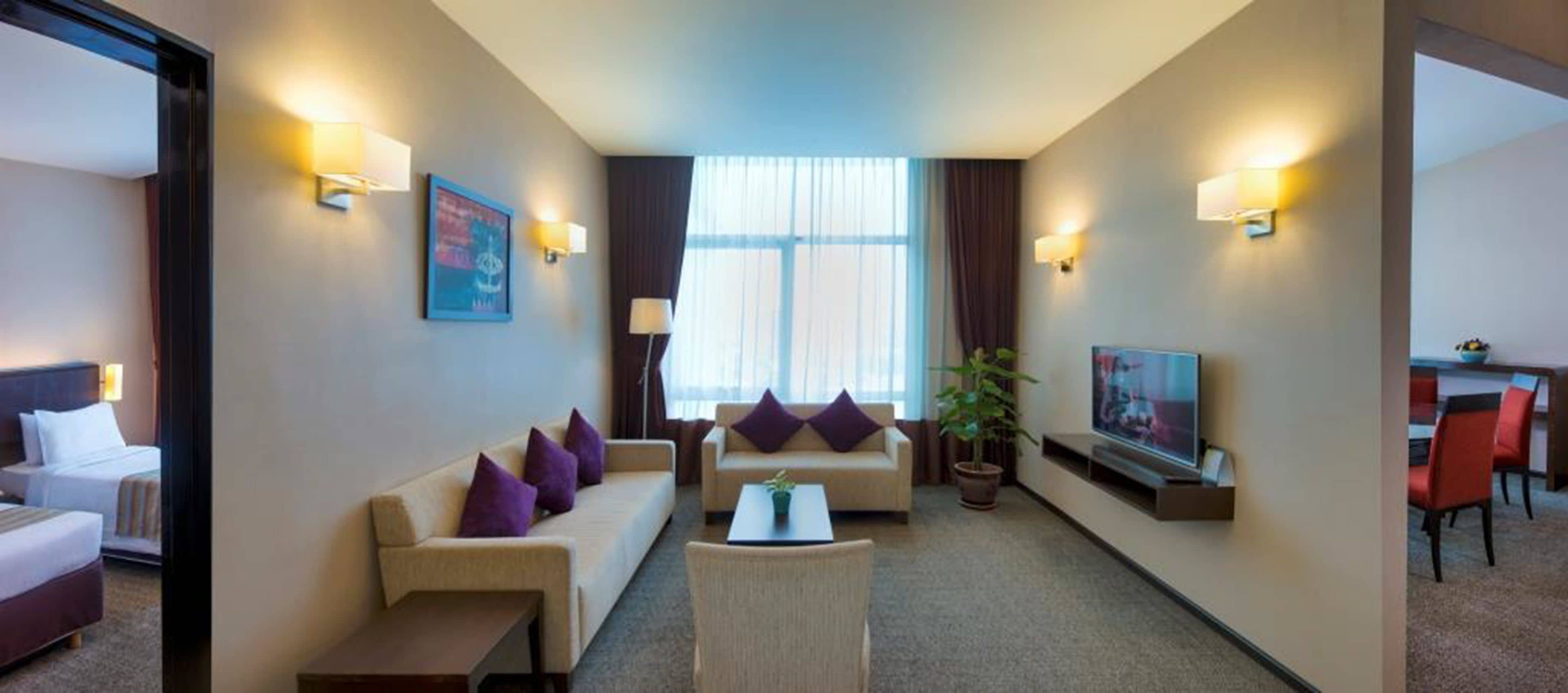 A hotel room with a bed, a sofa, a coffee table, a TV, and a dining area with chairs.