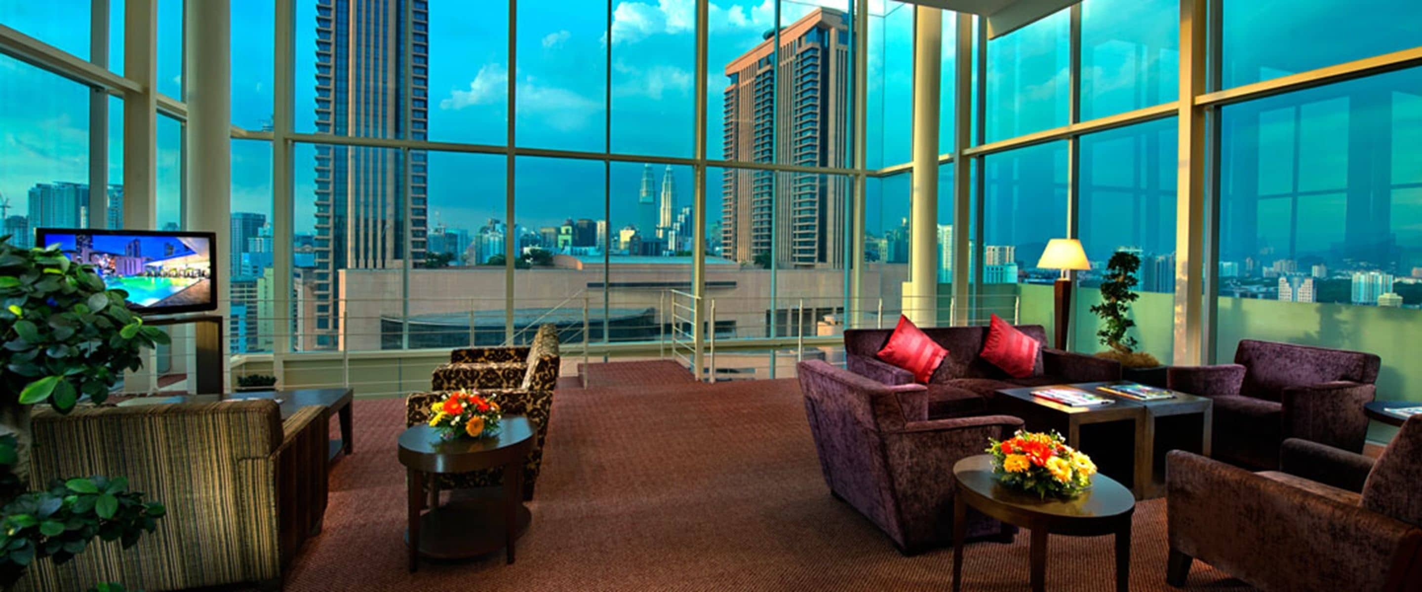 A high-rise hotel room with large windows offering a city view, a sofa, a coffee table, and a TV.