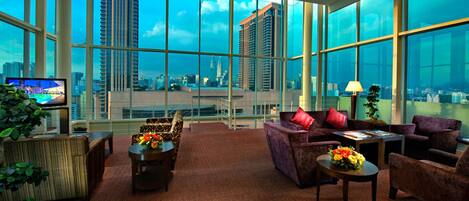 A high-rise hotel room with large windows offering a city view, a sofa, a coffee table, and a TV.