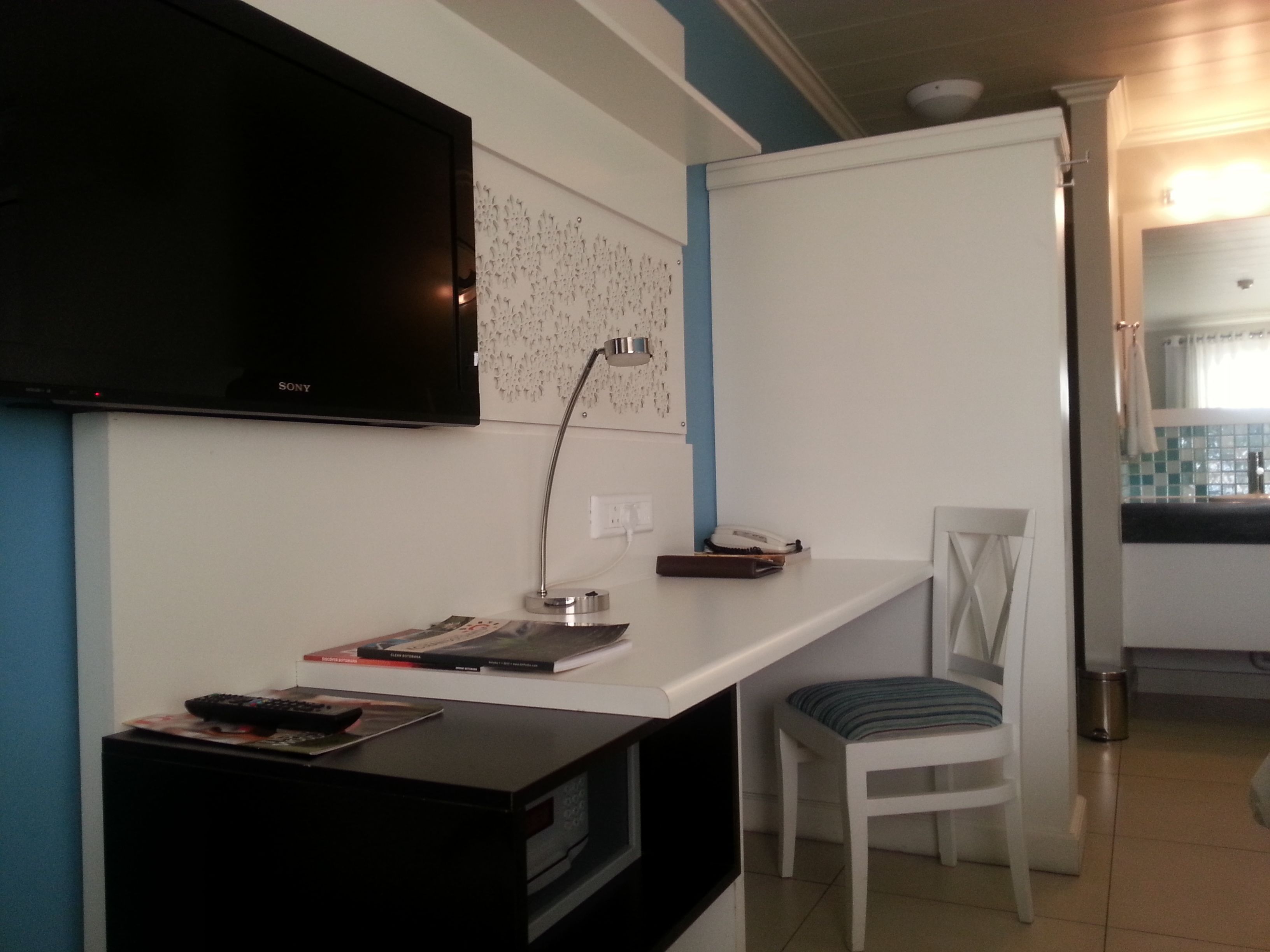 Un espace de vie compact comprenant un téléviseur à écran plat fixé au mur, un bureau avec une lampe, une chaise et un petit coin cuisine.
