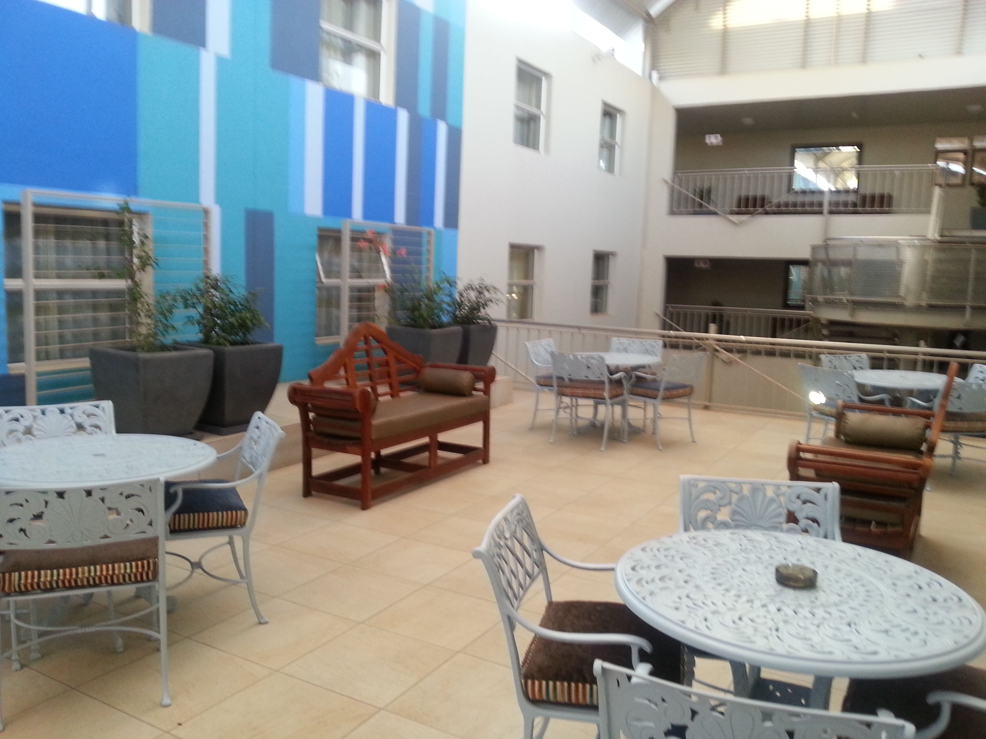 Un'area patio esterna con tavoli e sedie rotonde, piante in vaso e un edificio blu e bianco sullo sfondo.