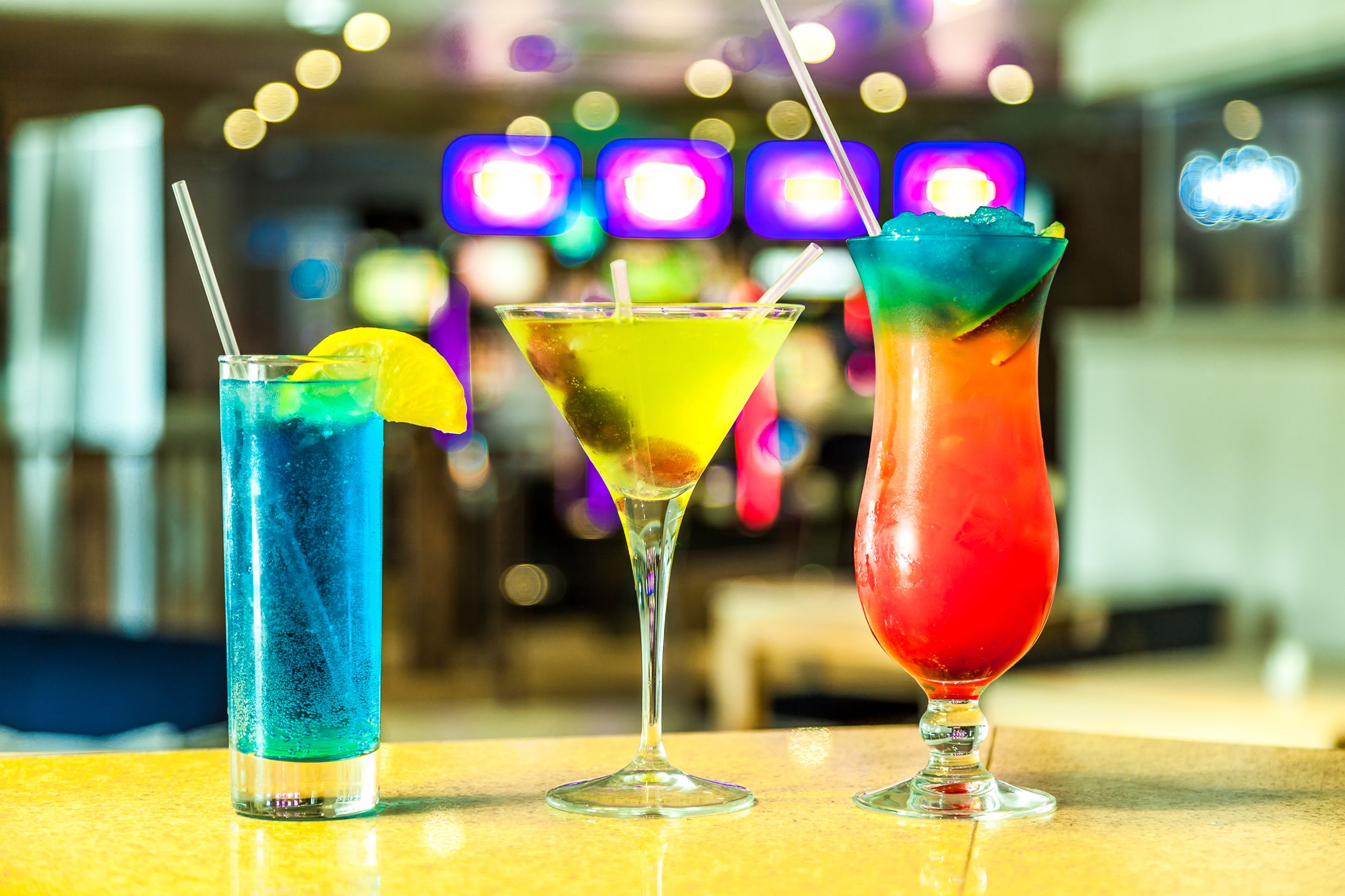 Three colorful cocktails on a bar counter with a blurred background of lights.