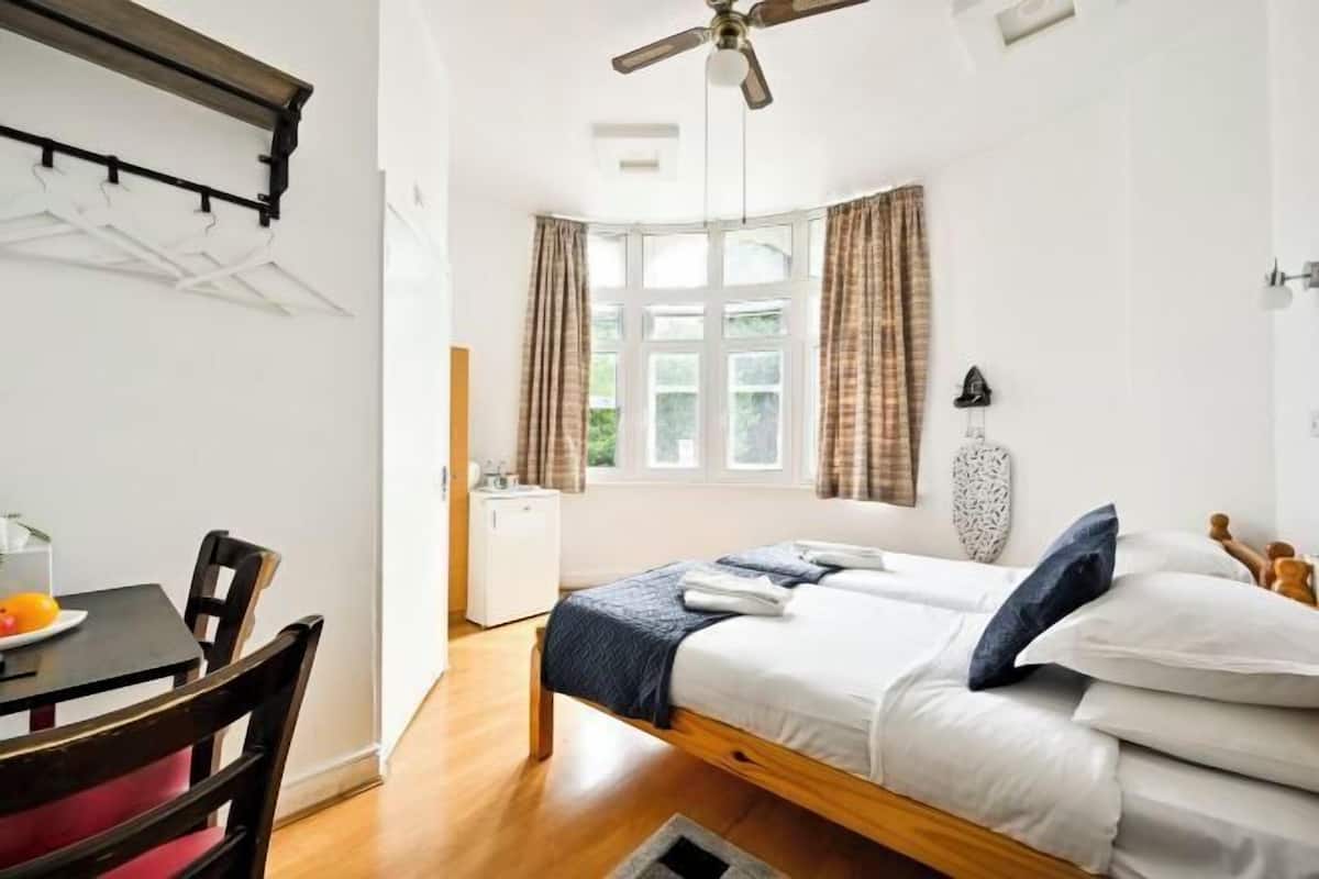 A bedroom with a bed, a desk, a chair, a ceiling fan, a window with curtains, and a small kitchenette.