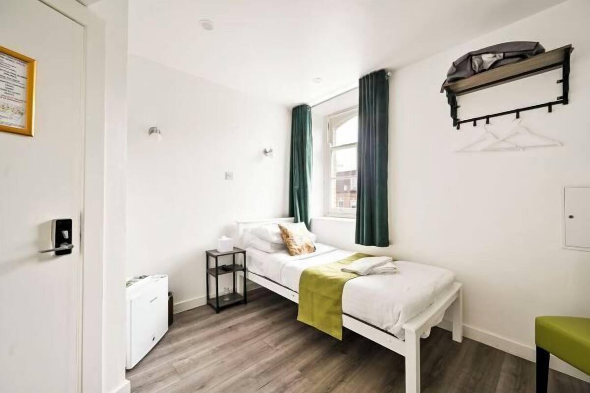 A single-bed room with a bedside table, a hanging coat rack, a window with curtains, and a small bench.