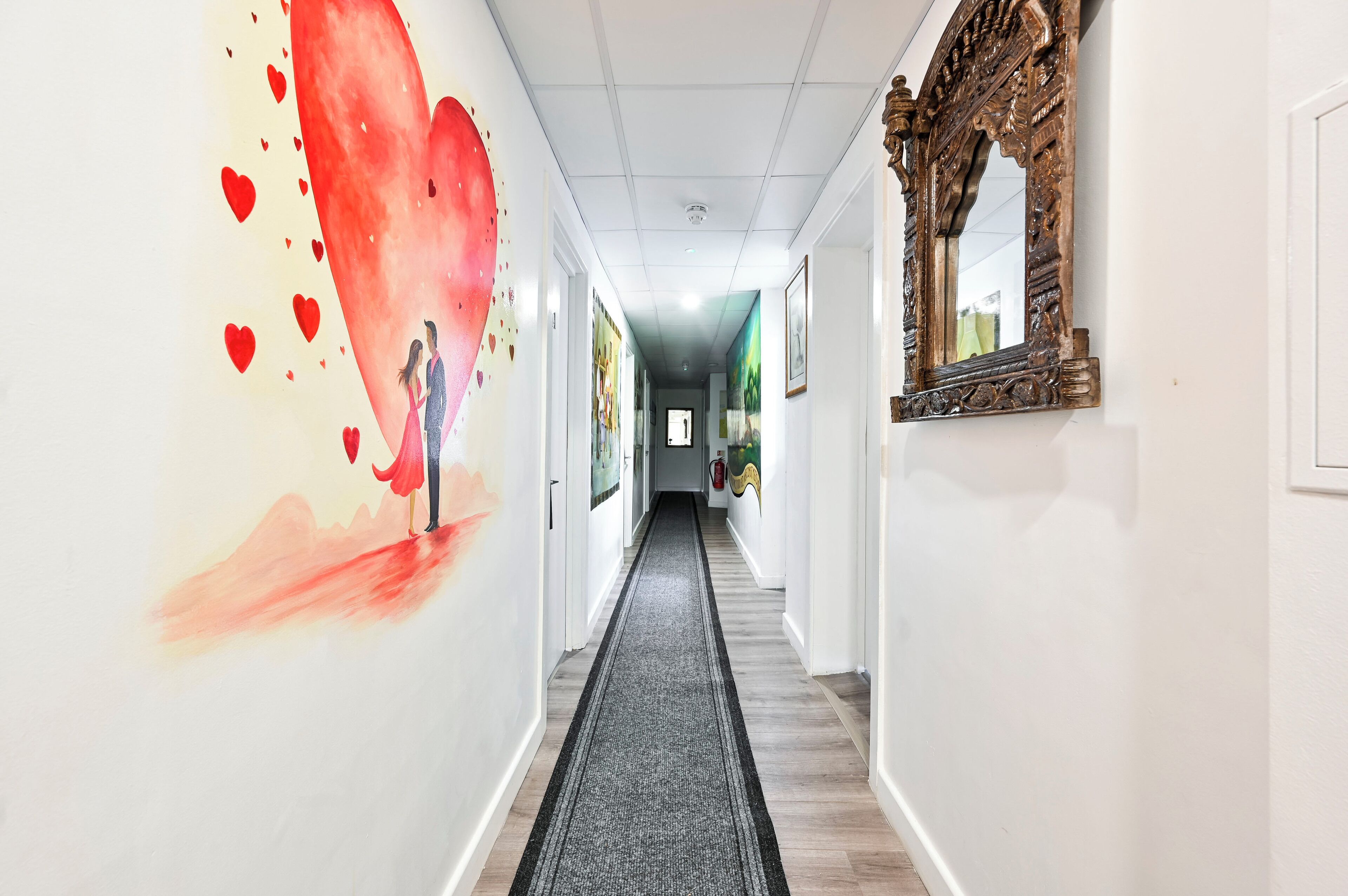 A hallway with a long carpet, a large wall mural, and a mirror.