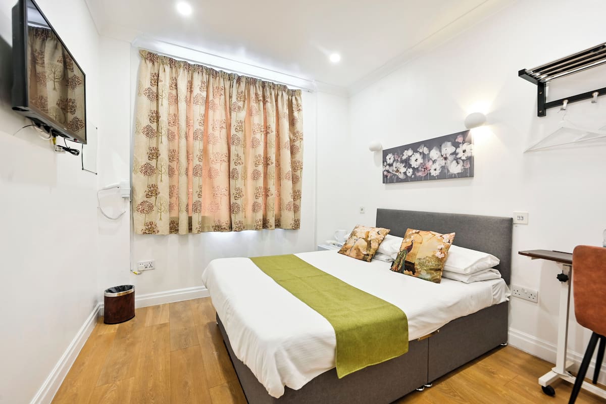 A bedroom with a bed, curtains, a painting, a TV, and a small trash bin.