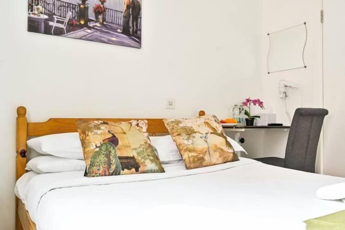A bed with patterned pillows, a bedside table with a vase of flowers, and a picture on the wall.
