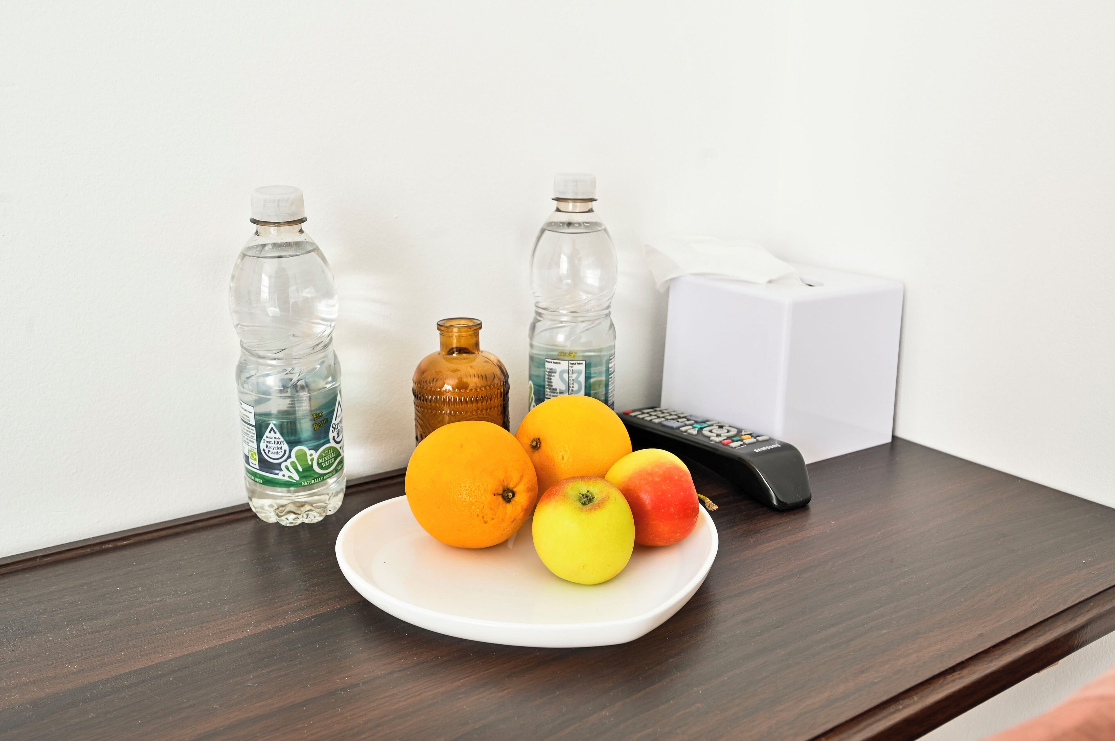 A plate with oranges and an apple on a wooden table, two water bottles, a remote control, and a tissue box.