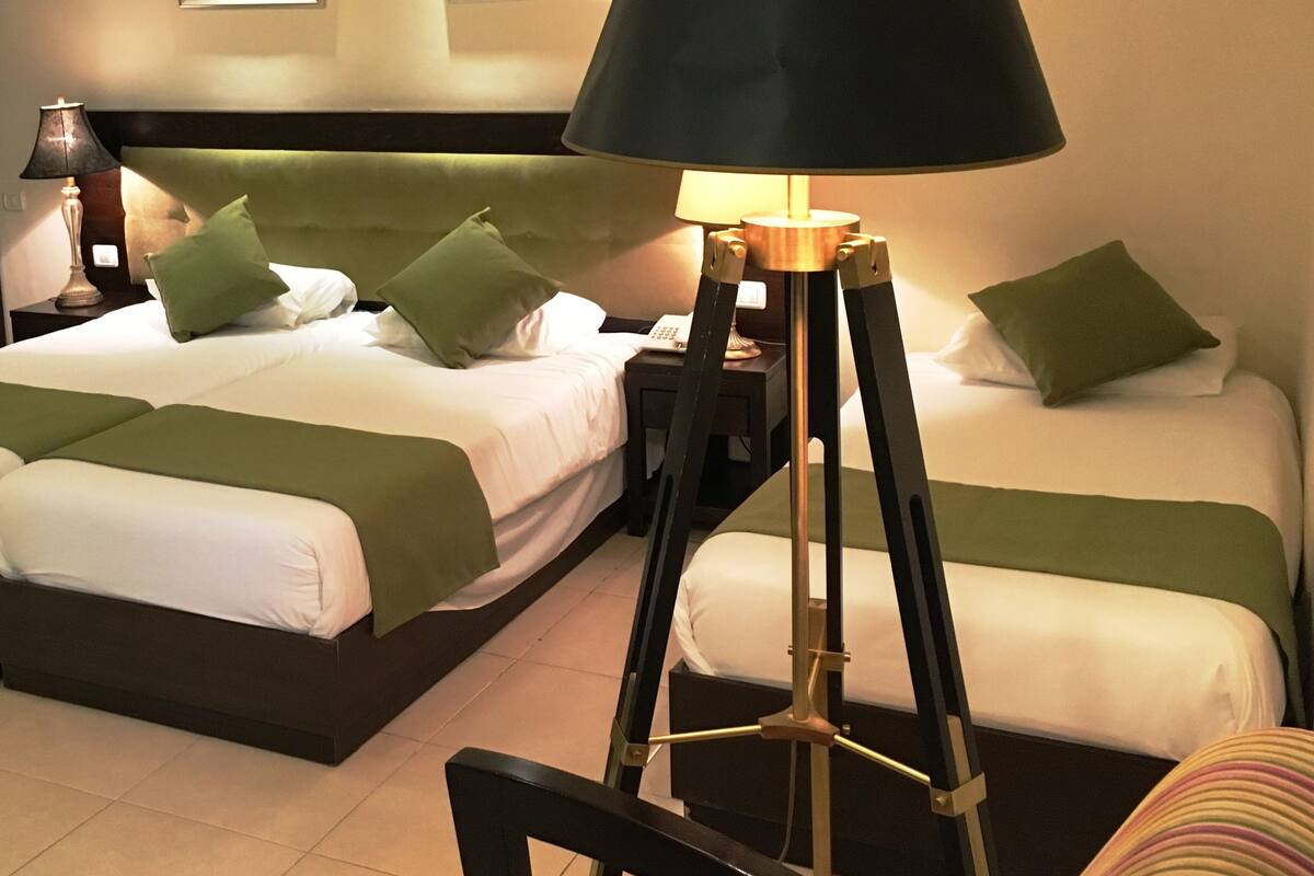 A hotel room with two beds, each with green and white bedding, a black floor lamp, and two framed pictures on the wall.