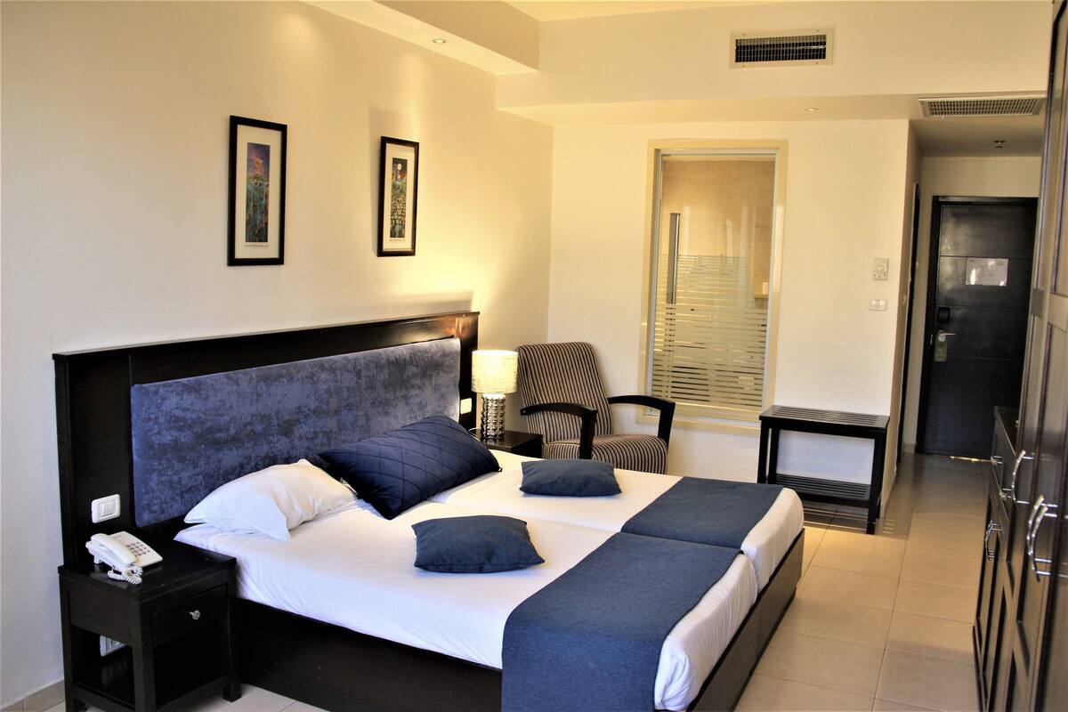 A hotel room with a large bed, a nightstand with a phone, a chair, and a desk with a lamp.