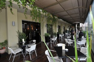 Un área de café al aire libre con mesas, sillas, plantas en macetas y un paseo techado.