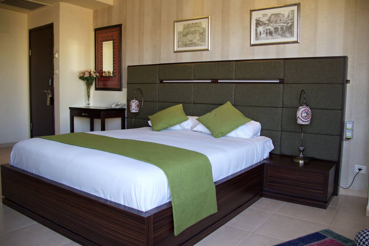 A large bed with a wooden frame, green pillows, and a white bedspread. There is a nightstand with a lamp and a vase with flowers.