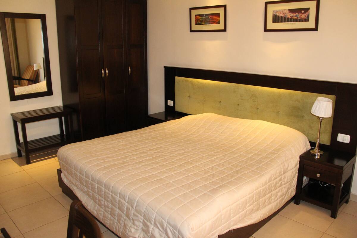 A hotel room with a large bed, a nightstand, a lamp, a wardrobe, and two framed pictures on the wall.