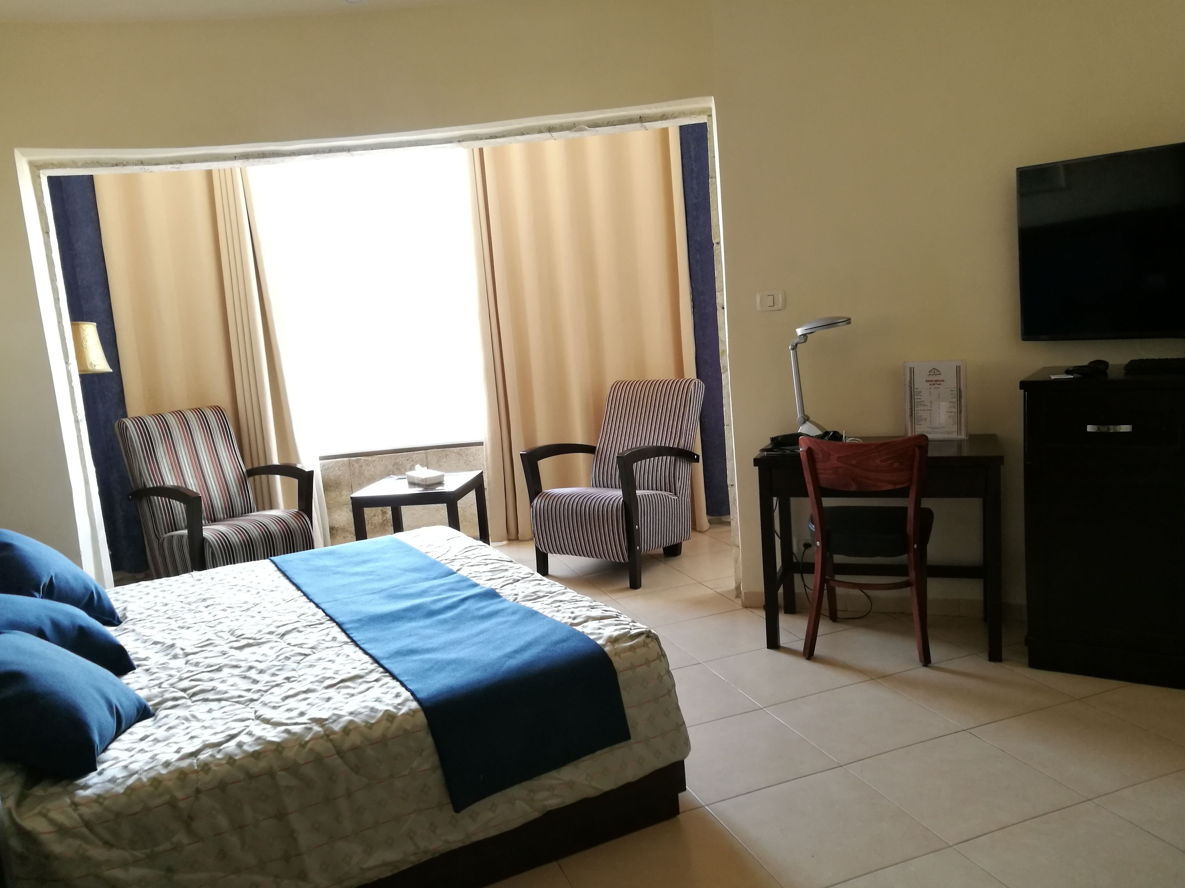 A hotel room with a bed, two striped armchairs, a desk with a lamp, a television, and a window with curtains.