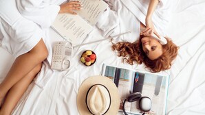 Uma pessoa deitada na cama com um livro, uma xícara, uma tigela de petiscos, um chapéu e fones de ouvido.