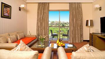 Quarto de hotel com sofá, mesa de centro com frutas e uma taça de vinho, televisão e varanda com vista para um jardim.