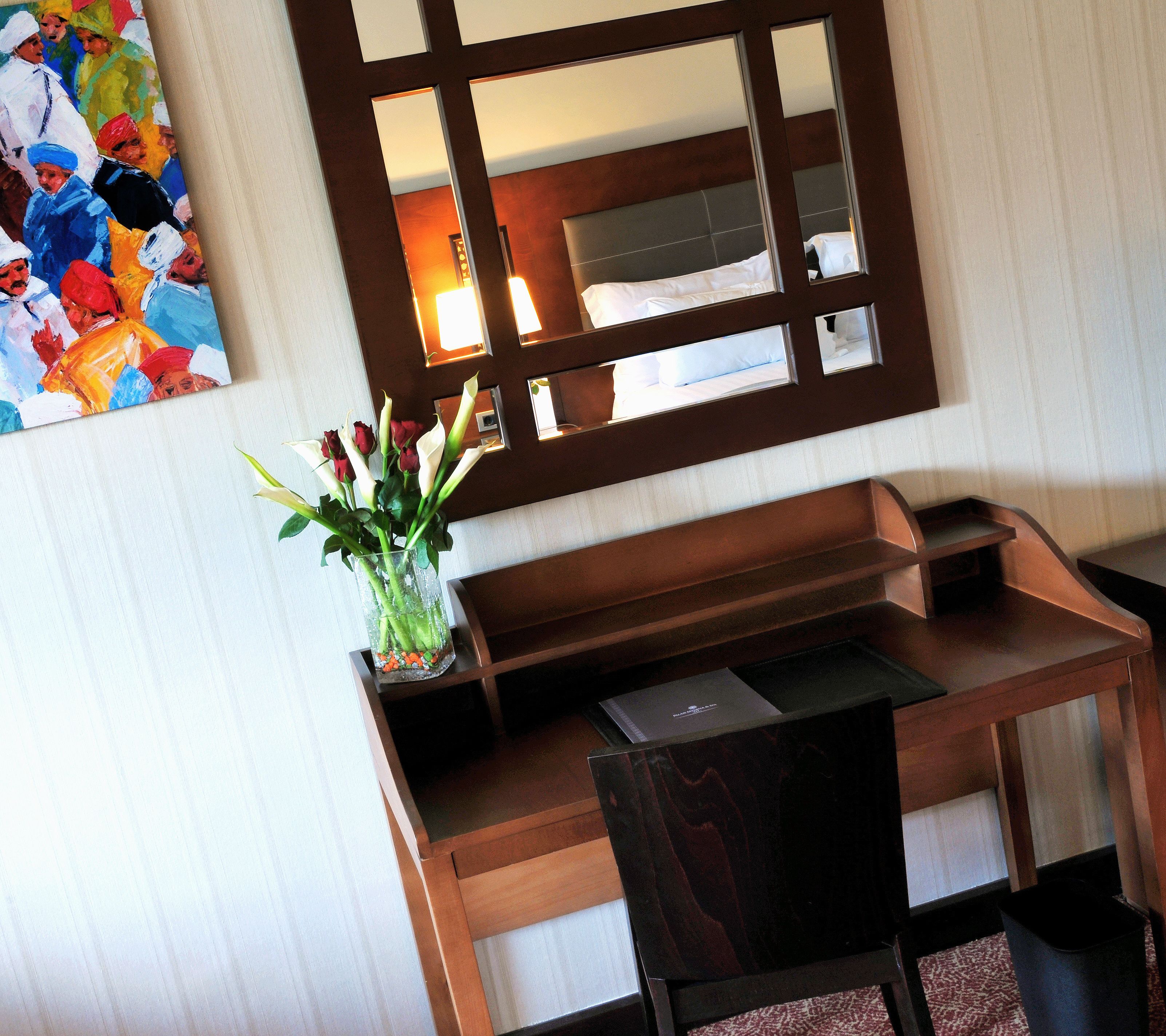 A hotel room with a wooden desk, a vase with flowers, a painting on the wall, and a large mirror above the bed.