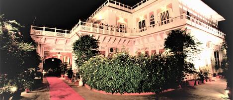 A large, illuminated building with a red carpet leading to an entrance, surrounded by potted plants and trees.