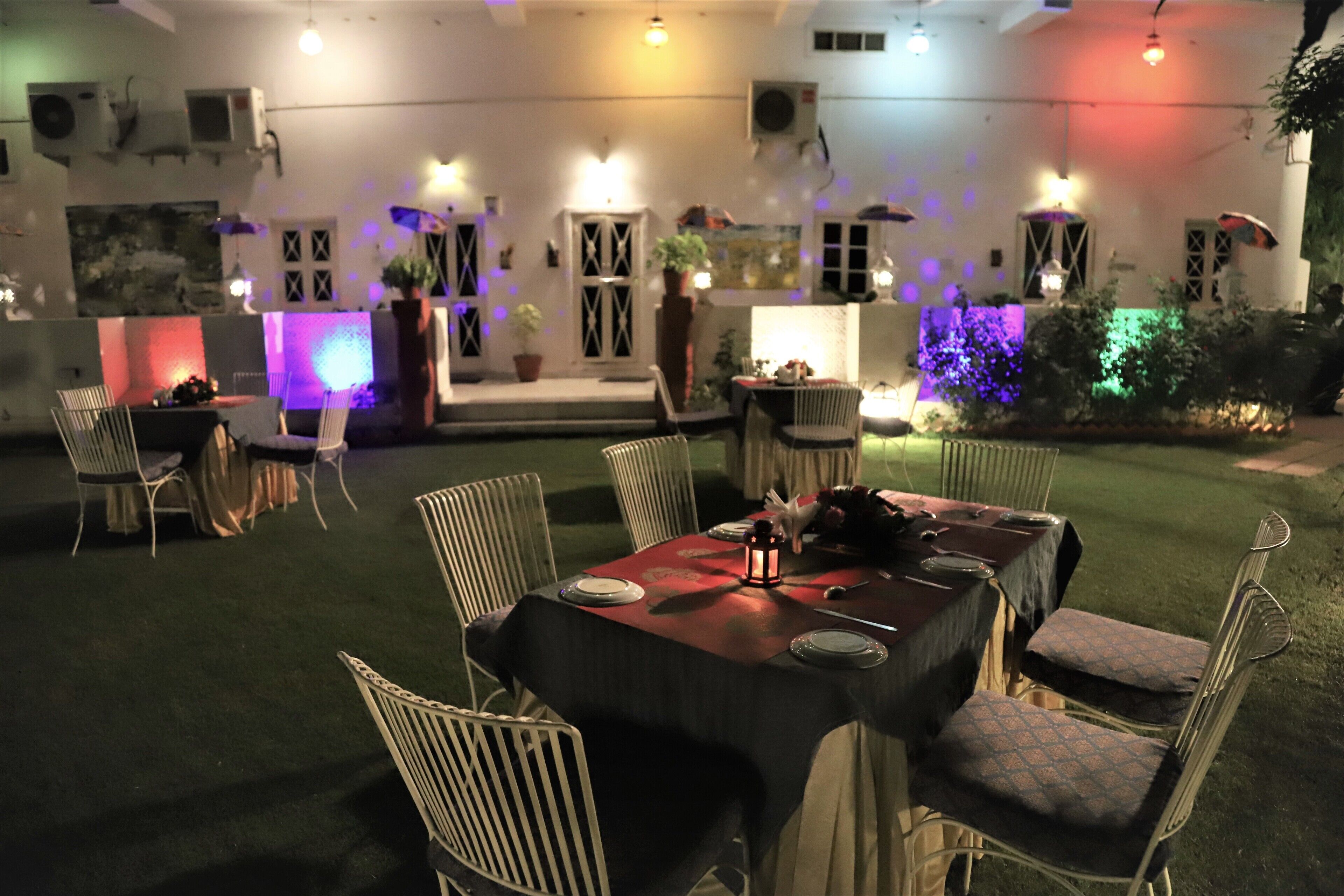 An outdoor dining area with tables and chairs, surrounded by a garden and illuminated by colorful lights.