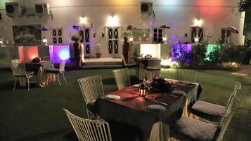An outdoor dining area with tables and chairs, surrounded by a garden and illuminated by colorful lights.