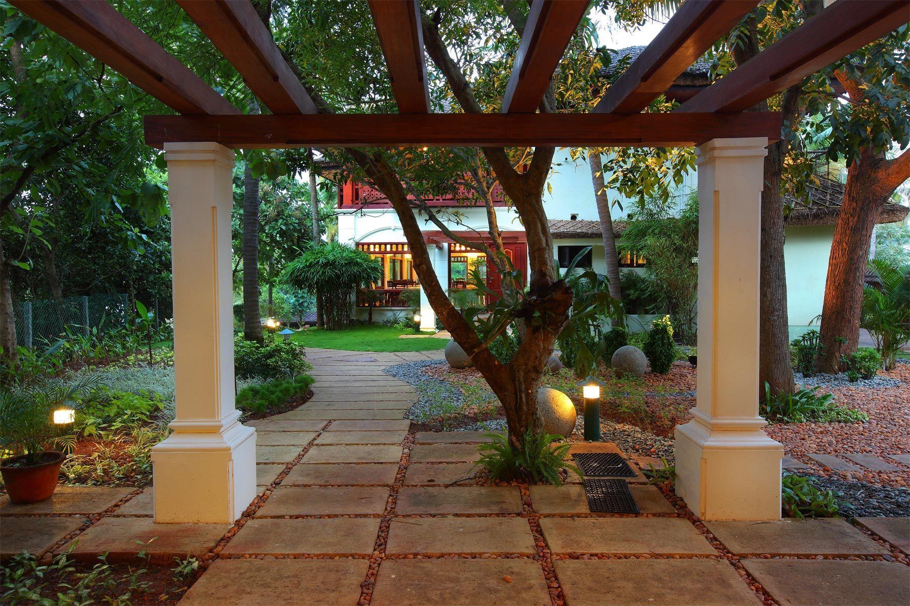Un chemin de pierres mène à une pergola en bois, d’où l’on aperçoit une maison au loin.