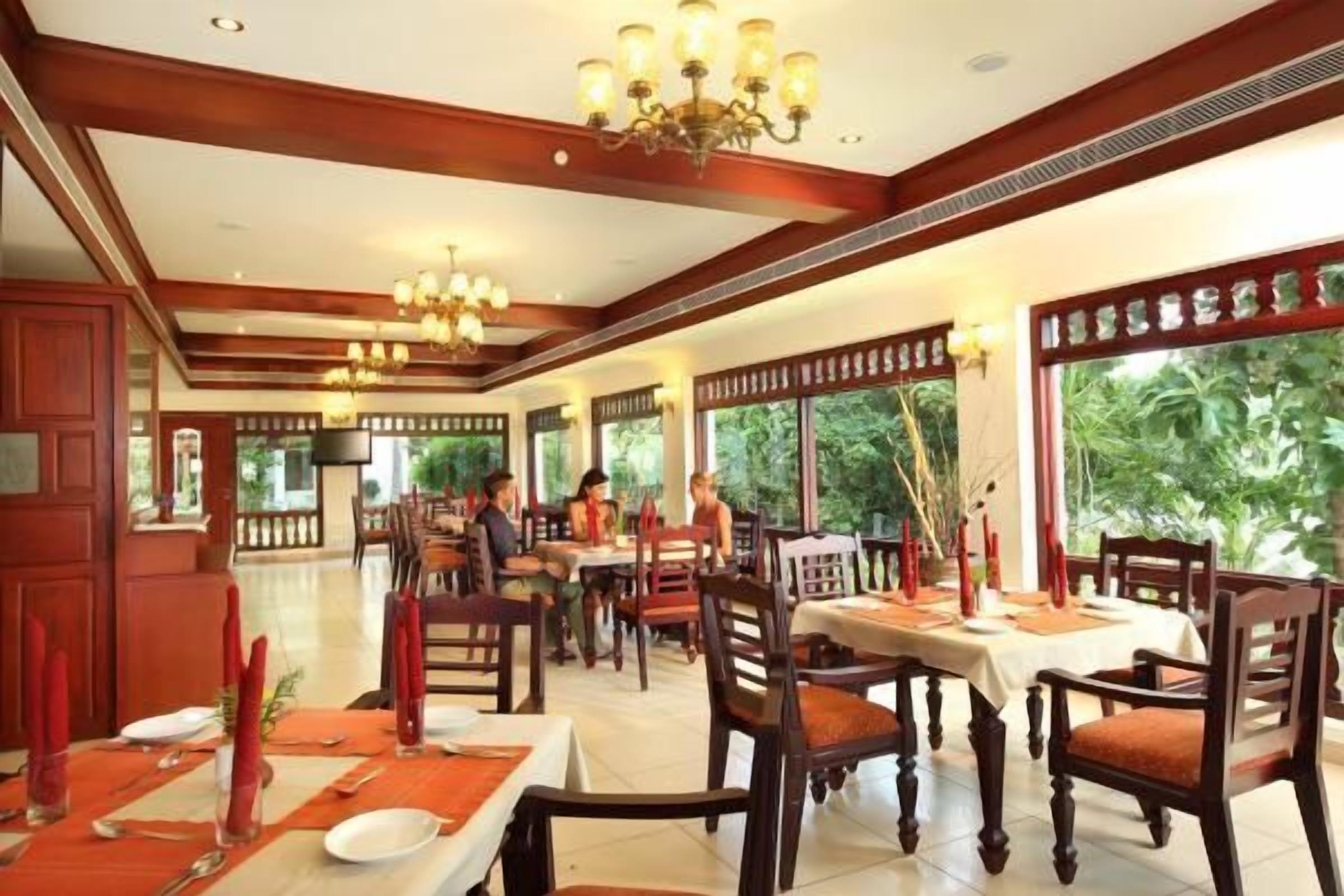 Un restaurante con mobiliario de madera, candelabros y grandes ventanales con follaje verde en el exterior.