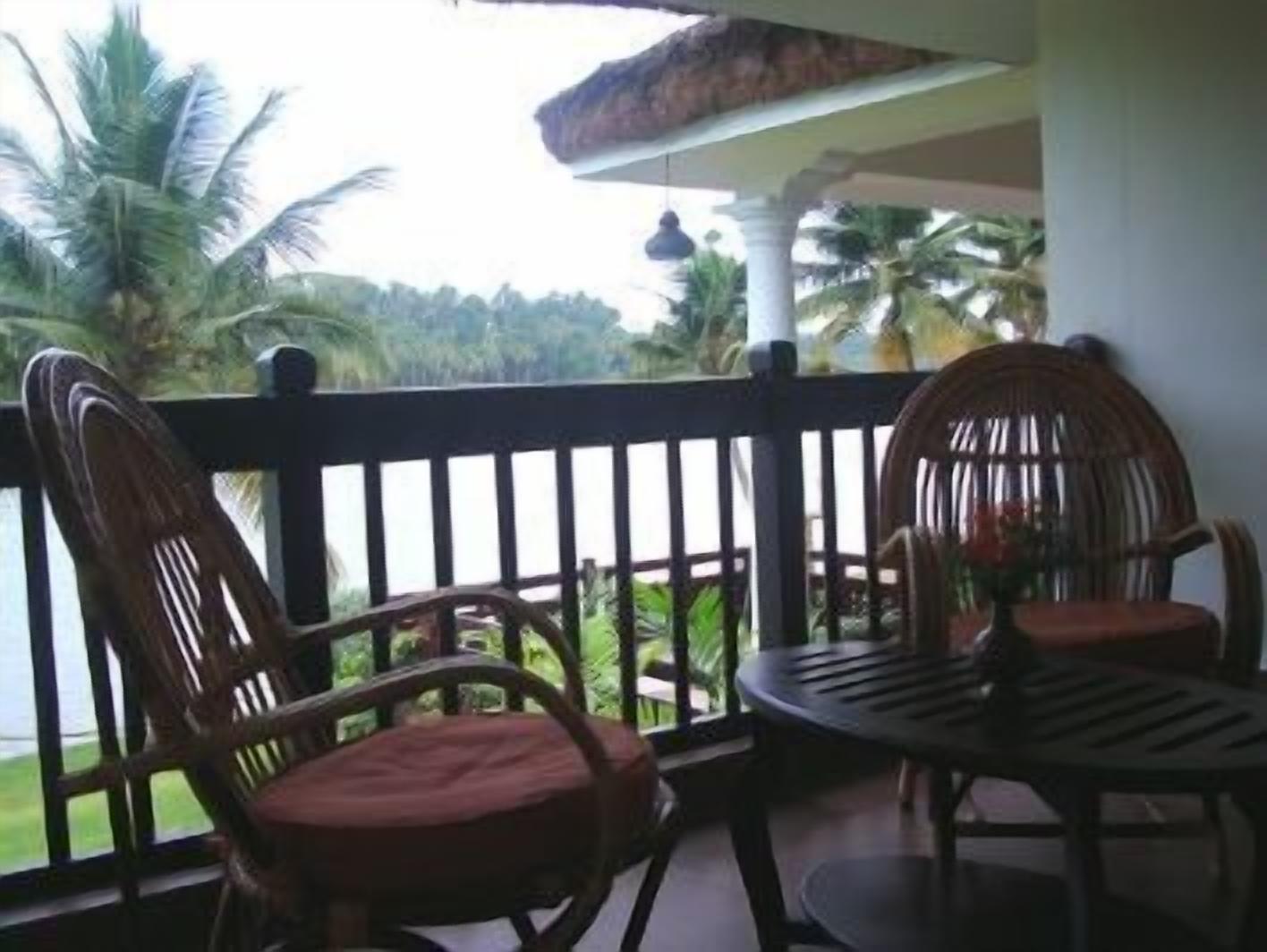 A balcony with two wicker chairs and a small table, overlooking a lush green landscape with palm trees.