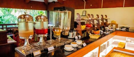 Zona de bufé con dispensadores de café, una cesta de frutas y diversos platos.