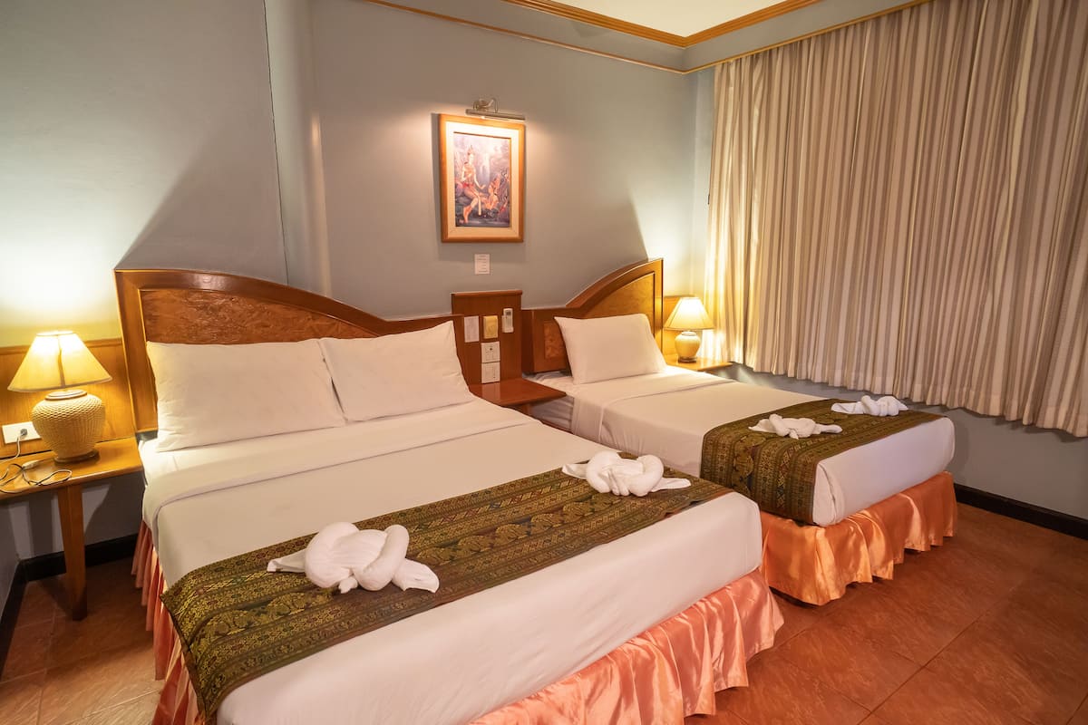 Quarto de hotel com duas camas, cada uma com cabeceira de madeira, roupa de cama branca e um edredom estampado em verde. Há duas luminárias de mesa, um quadro na parede e uma janela com cortinas.